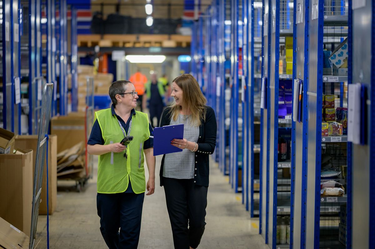 Two people talking as they walk down a warehouse aisle lined with stocked shelving.