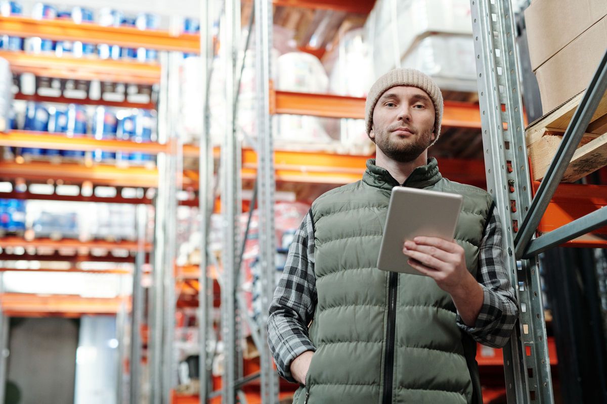 A man in a puffer vest holding a tablet between rows of pallet racking in a warehouse.